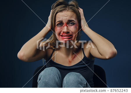 Scared woman, bleeding face covered with bruises, in black t-shirt and jeans, grabbed own head and crying, sitting on blue background. Close-up Scared woman, bleeding face covered with bruises, in black t-shirt and jeans, grabbed own head and crying, sitting on blue background. Close-up 87652243