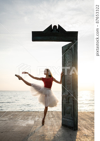 Female ballet dancer is posing near art object on seafront during sunrise 87653322