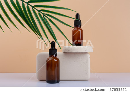 Two dark transparent glass bottles with skin serum or oil for manicure on gypsum podium against beige background with palm leaf Two dark transparent glass bottles with skin serum or oil for manicure on gypsum podium against beige background with palm leaf 87655851