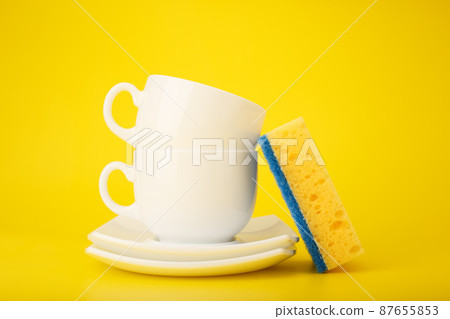 Dishwashing minimal concept. Plates, cups and cleaning sponge against bright yellow background Dishwashing minimal concept. Plates, cups and cleaning sponge against bright yellow background 87655853