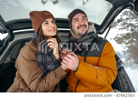 Lovely smiling couple sitting in car trunk in winter forest Lovely smiling couple sitting in car trunk in winter forest 87662565
