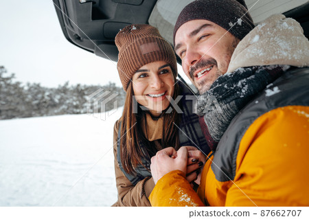 Lovely smiling couple sitting in car trunk in winter forest Lovely smiling couple sitting in car trunk in winter forest 87662707