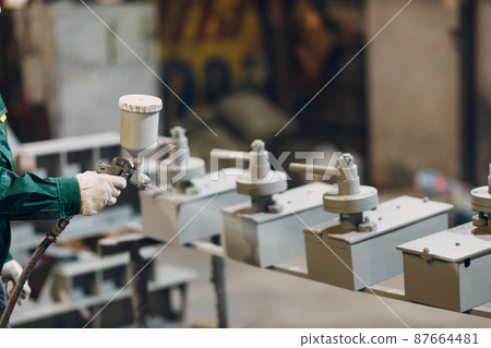 Powder primer coating of metal parts. Worker man in a protective suit sprays powder paint from gun on metal product construction at factory plant. 87664481