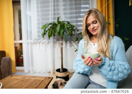 An attractive blonde is sitting on the sofa with a mug of coffee. 87667026