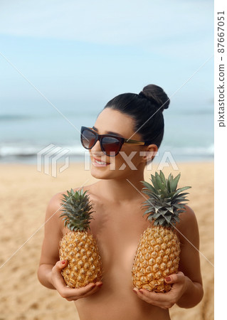 Beautiful caucasian sunbathed woman model with dark swimsuit posing on summer beach on blue sky and ocean background. Covering  her breast with pineapples 87667051