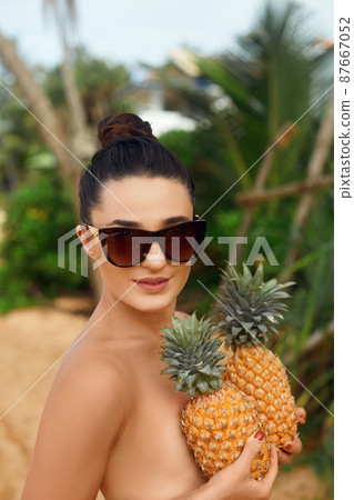 Beautiful cheerful nude girl holding a pineapple at the breast on the beach. Woman with bronze tan in bikini. Covering her breast with pineapples 87667052