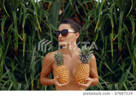 Beautiful cheerful nude girl holding a pineapple at the breast on the tropical background . Woman with bronze tan in bikini. Covering her breast with pineapples 87667224