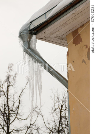 Frozen ice icicles hang from a drainpipe at the edge of the roof. Frozen ice icicles hang from a drainpipe at the edge of the roof. 87668282