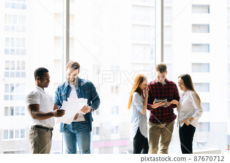 Front view of team young multiracial startup business men and women discussing together new project, reading documents and using digital tablet, standing on background of window and skyscrapers. Front view of team young multiracial startup business men and women discussing together new project, reading documents and using digital tablet, standing on background of window and skyscrapers. 87670712