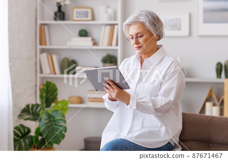 Focused mature woman using tablet at home 87671467