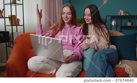 Happy two female women couple family on sofa at home, enjoy chatting, spend leisure time on laptop 87674640