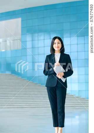 一位身著年輕西裝的女商人站在辦公室入口處拿著巴索松 一位身著年輕西裝的女商人站在辦公室入口處拿著巴索松 87677480