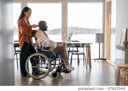Young woman with electric trimmer cutting hair on head of man with disability 87681949