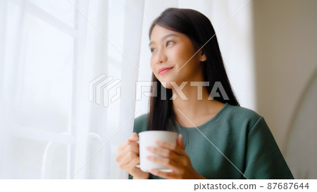 Young asian woman standing beside window and holding mug in bedroom at home 87687644