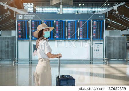 Young female wearing face mask with luggage walking in airport, protection Coronavirus disease infection, Asian woman traveler with hat. Time to travel, Test and Go and vaccine booster dose concept 87689859