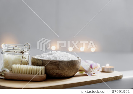 Preparation for hotel spa treatment or home bath procedure. White washbasin in bathroom with accessories on tray. Burning candles, washcloth, soap, foot brush, bottle with sea salt. Blurred background 87691961