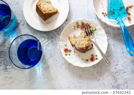 Two slices of banana cakes on white plate with fork and blue glasses of water on concrete table top in top view. 87692342