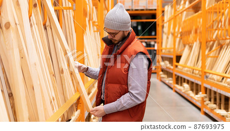 a male builder in a store chooses lumber among wooden bars and planks a male builder in a store chooses lumber among wooden bars and planks 87693975