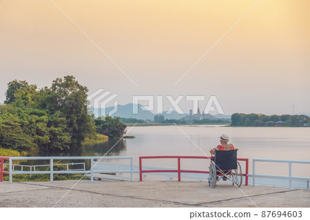 Back view of Asian elderly female patient wearing surgical mask and sitting on wheelchair with feeling lonely and depressed at riverside in the evening with view of Wat Tham Sua in background. Back view of Asian elderly female patient wearing surgical mask and sitting on wheelchair with feeling lonely and depressed at riverside in the evening with view of Wat Tham Sua in background. 87694603