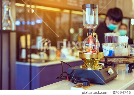 Close up to barista make coffee with Vacuum Pot Syphon Brewer. Syphon (Siphon) coffee maker brews drink using two chambers where vapor pressure and vacuum produce coffee in cafe. Selective focus. Close up to barista make coffee with Vacuum Pot Syphon Brewer. Syphon (Siphon) coffee maker brews drink using two chambers where vapor pressure and vacuum produce coffee in cafe. Selective focus. 87694746