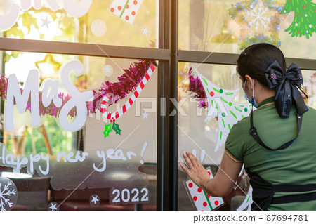 Asian woman decorates beautiful stickers on window glass in front of cafe to welcome Christmas and New Year. Prepare to decorate the storefront to be beautiful during the important holidays of year. 87694781