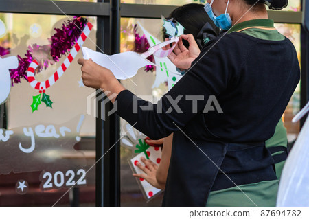Asian women decorate beautiful stickers on window glass in front of cafe to welcome Christmas and New Year. Prepare to decorate the storefront to be beautiful during the important holidays of year. 87694782