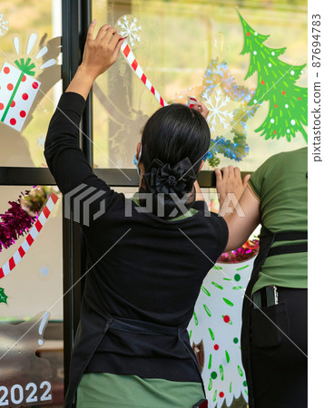Asian women decorate beautiful stickers on window glass in front of cafe to welcome Christmas and New Year. Prepare to decorate the storefront to be beautiful during the important holidays of year. 87694783
