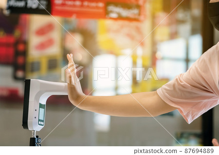Customer with protective mask raised hand at digital thermometer with temperature scanning machine for fever before entering area. Screening to prevent infected people from entering restricted areas. Customer with protective mask raised hand at digital thermometer with temperature scanning machine for fever before entering area. Screening to prevent infected people from entering restricted areas. 87694889