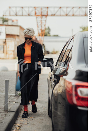 Stylish Young Woman in Jacket and Sunglasses Moving Near the Black Car 87697573