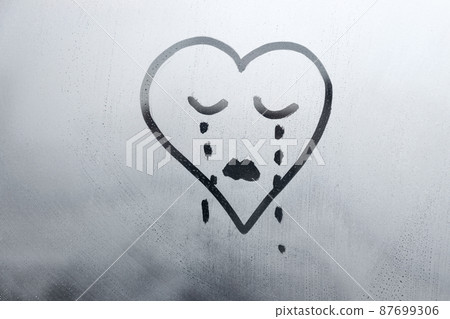 drawn sad face in shape of heart with closed eyes and tears on foggy glass window raindrops blue color concept photo 87699306