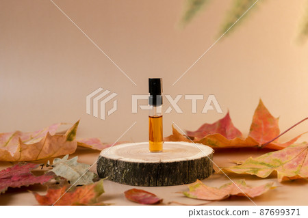 Glass perfume sample with brown liquid on a wooden tray standing on beige background with fallen leaves around. Luxury and natural autumn cosmetics presentation. Tester on a woodcut. Front view 87699371