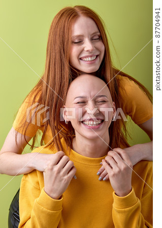 two Joyful young bald and redhead women hugging posing isolated on green background two Joyful young bald and redhead women hugging posing isolated on green background 87704941