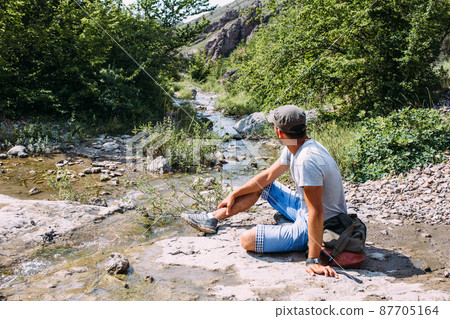 Young man traveler walking alone by a mountain river with rocky summer landscape Young man traveler walking alone by a mountain river with rocky summer landscape 87705164