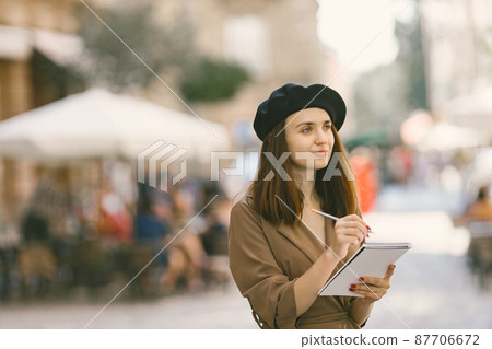 Girl in dress and black hat painting a sketch while standing on a street 87706672