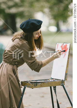 Girl in dress and black hat painting on an easel in a park 87706724