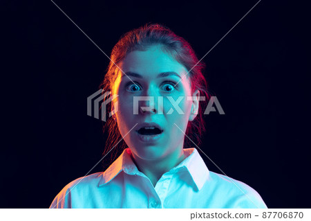 Shocked young girl in white shirt looking at camera with open mouth isolated on dark background in blue neon light, filter. Concept of emotions, fashion, youth Shocked young girl in white shirt looking at camera with open mouth isolated on dark background in blue neon light, filter. Concept of emotions, fashion, youth 87706870