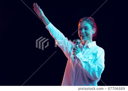 Half-length portrait of young pretty girl in white shirt with microphone isolated on dark background in blue neon light, filter. Concept of emotions, fashion, youth Half-length portrait of young pretty girl in white shirt with microphone isolated on dark background in blue neon light, filter. Concept of emotions, fashion, youth 87706889