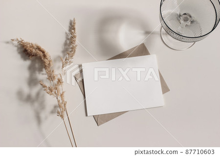 Feminine stationery still life. Champagne, cocktail glass, shadows. Blank greeting card, invitation mockup, craft envelope. Dry grass, festuca plant in sunlight. Beige table background. Fall flatlay 87710603