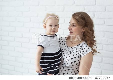 Mother holds her child with hearing aids and cochlear implants on white brick wall with copy space and place for advertising . Deaf and health concept 87712057