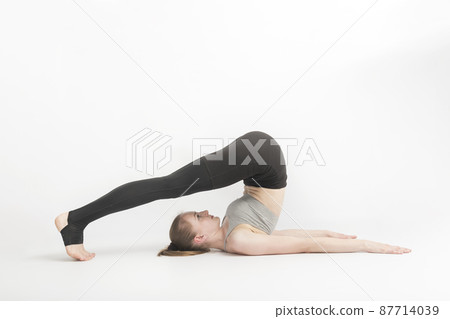 Halasana. Plow Pose. Young woman during a yoga class. Side view. Yoga trainer shows technique of performing asana on white background. Halasana. Plow Pose. Young woman during a yoga class. Side view. Yoga trainer shows technique of performing asana on white background. 87714039