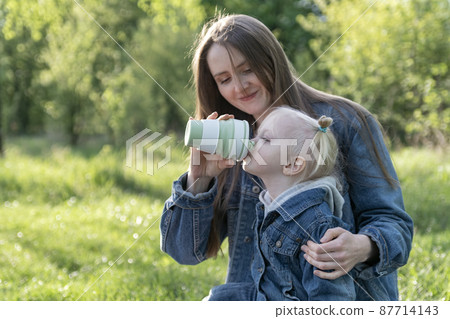 Young mother and little daughter in the spring against on green park background. Mom drinks her daughter from silicone cup. Young mother and little daughter in the spring against on green park background. Mom drinks her daughter from silicone cup. 87714143