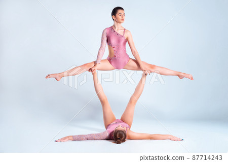 Two flexible girls gymnasts with pigtails, in pink leotards are performing splits using support while posing isolated on white background. Close-up. 87714243