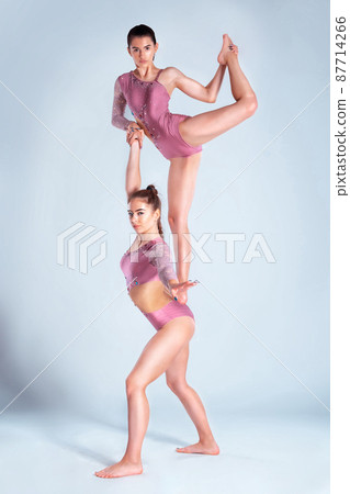 Two flexible girls gymnasts in beige leotards performing complex elements of gymnastics using support, posing isolated on white background. Close-up. Two flexible girls gymnasts in beige leotards performing complex elements of gymnastics using support, posing isolated on white background. Close-up. 87714266