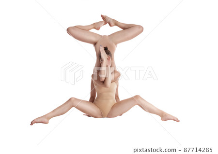Two flexible girls gymnasts in beige leotards performing complex elements of gymnastics using support, posing isolated on white background. Close-up. Two flexible girls gymnasts in beige leotards performing complex elements of gymnastics using support, posing isolated on white background. Close-up. 87714285