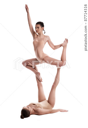 Two flexible girls gymnasts in beige leotards are performing exercises upside down using support and posing isolated on white background. Close-up. 87714316