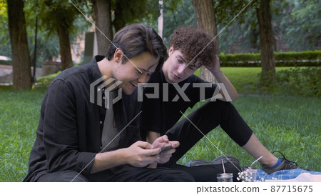 The guy plays the game on the phone, and the second watches the process in nature in the summer. LGBT The guy plays the game on the phone, and the second watches the process in nature in the summer. LGBT 87715675
