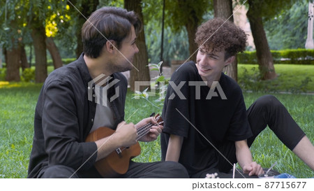 A guy plays the ukulele to another guy in nature in the summer. LGBT A guy plays the ukulele to another guy in nature in the summer. LGBT 87715677