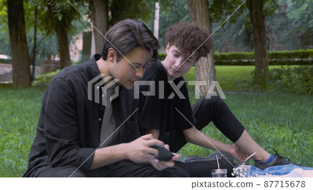 The guy plays the game on the phone, and the second watches the process in nature in the summer. LGBT The guy plays the game on the phone, and the second watches the process in nature in the summer. LGBT 87715678