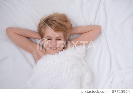 A beautiful teenage girl is lying in bed. Happy child smiles. The concept of good sleep and pleasant awakening. White fluffy blanket. There is space for text. Sincere emotions 87715904