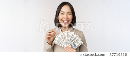 Image of successful businesswoman holding money. Asian corporate woman with cash dollars, smiling and laughing, standing over white background Image of successful businesswoman holding money. Asian corporate woman with cash dollars, smiling and laughing, standing over white background 87719316